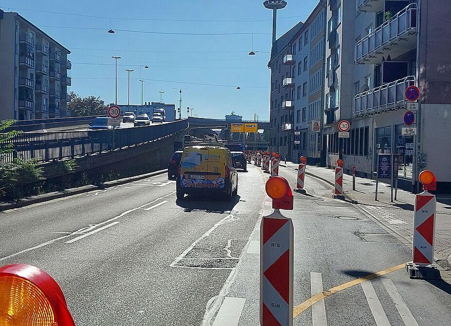 Luisenring: Gesperrte Fahrspur statt Radweg Blick entlang des Luisenrings in Mannheim bei Sonnenschein. Links fahren Autos und ein Lieferwagen in Richtung Hochstraße. Die rechte Fahrspur ist mit einer Reihe rot-weißer Warnbaken mit gelben Warnleuchten und einer gelben Linie abgesperrt. Darunter sind noch alte weiße Fahrbahnmarkierungen zu sehen.