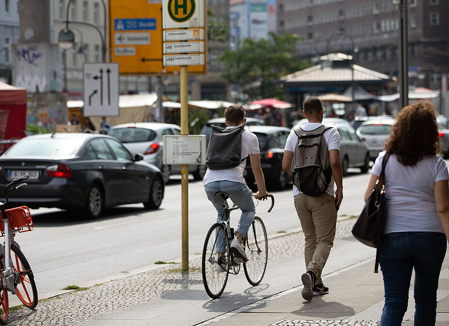 Geteilter Verkehrsraum in der Stadt Eine Straßenszene: Ein Fahrradfahrer und zwei Fußgänger teilen sich einen gepflasterten Gehweg. Im Hintergrund parken Autos am Straßenrand.