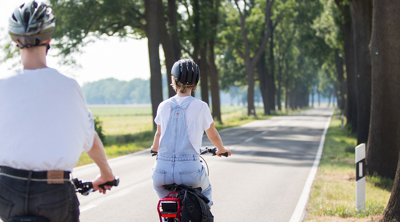 Zwei Personen mit Fahrradhelmen fahren auf einer schmalen Landstraße ohne separaten Radweg.